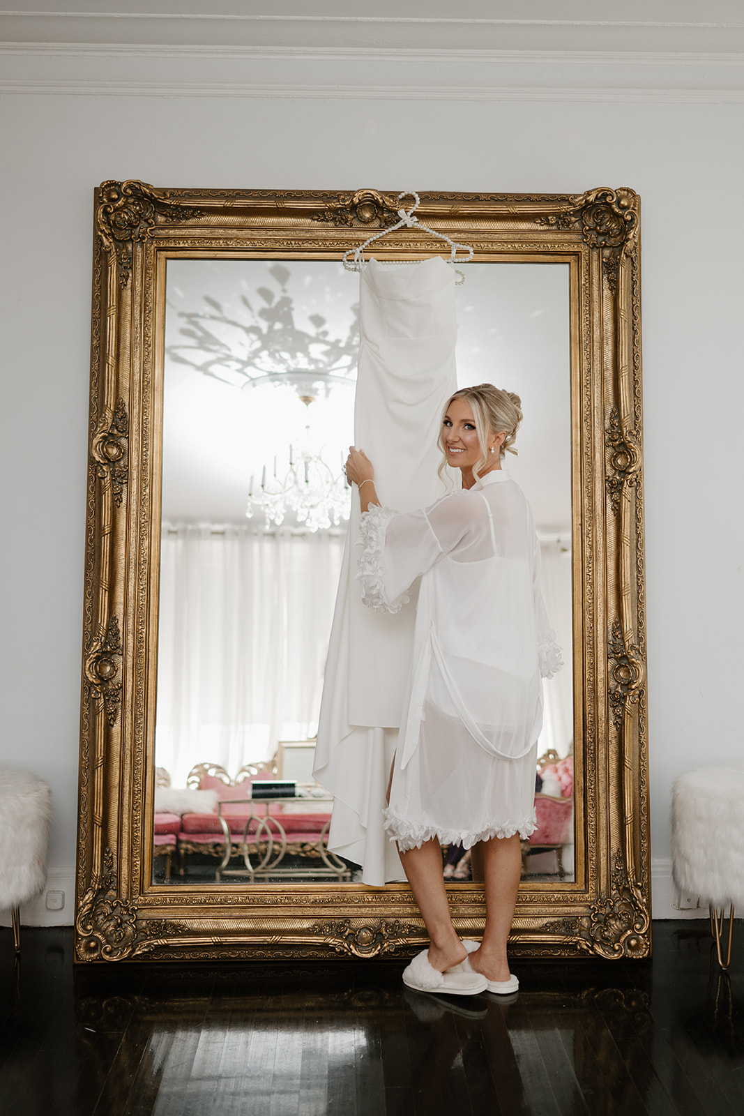 Bride in a white lace robe holds her wedding gown in front of an ornate gold mirror, smiling with anticipation.